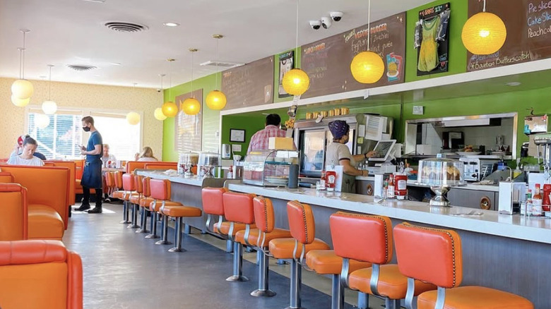 Bright orange booths and counter stools lined up at a diner counter. The diner has green walls and yellow globe lamps.