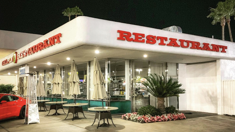 The exterior of a sleek, shiny restaurant at night with a bright red sportscar and tables with umbrellas outside