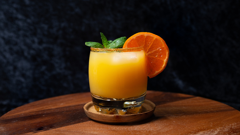 Whiskey cocktail featuring orange juice with fresh orange slice and mint on wood table.
