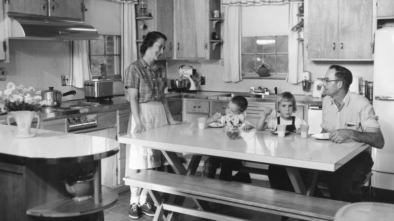 A woman smiles at her family eating in the kitchen at a table