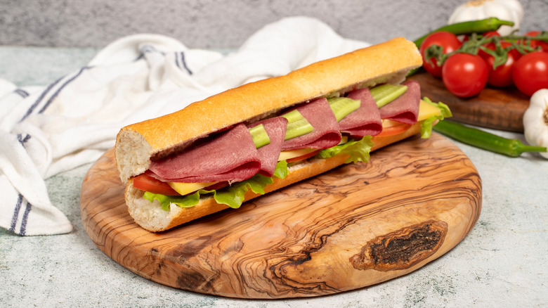 Sub sandwich with sliced beef tongue, lettuce, cheese, pickles, and more on a wood tray with tomatoes on the vine to the right and a dishtowel in back