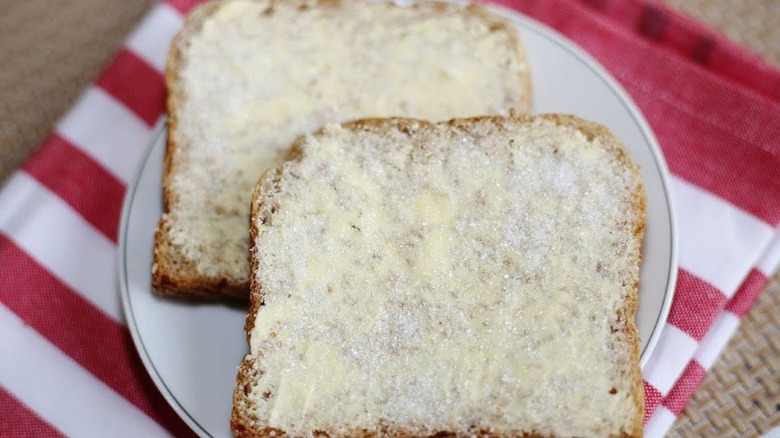 Two slices of white bread topped with butter and sugar on a white plate and red and white striped kitchen towel