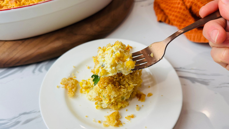 Hand holding forkful of scalloped corn over plate with larger serving