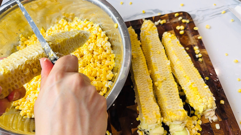 Knife scraping along corn cob over bowl next to cobs on wooden board