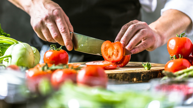A hand slicing fresh tomatoes with other vegetables around them