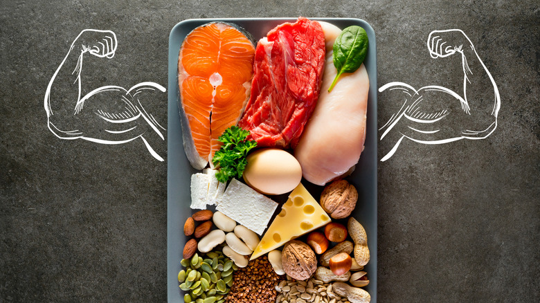 Tray of high-protein foods, including salmon, beef, chicken, and cheese, on a slate background with muscled arms drawn with chalk on either side