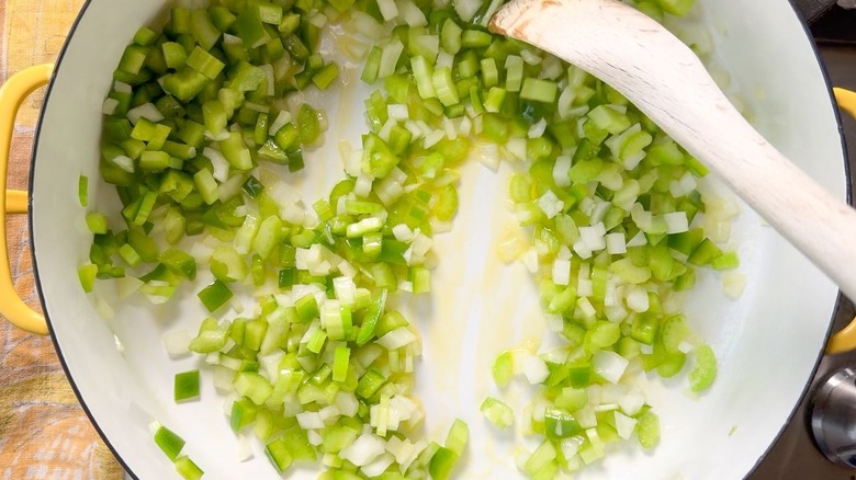 Stirring chopped onion, green bell pepper, and celery in olive oil with wooden spoon in large yellow pot on stovetop