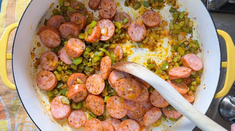Stirring sliced Andouille sausage and vegetables with wooden spoon in large yellow pot