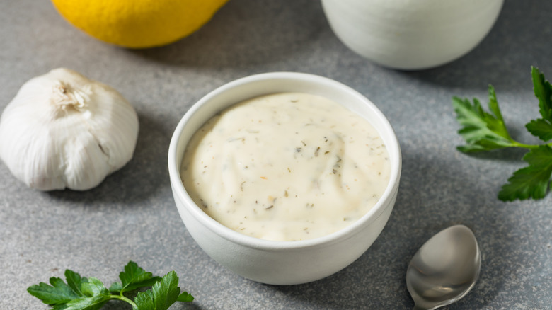 A bowl of ranch dressing on a table with a lemon, garlic, and herbs in the background