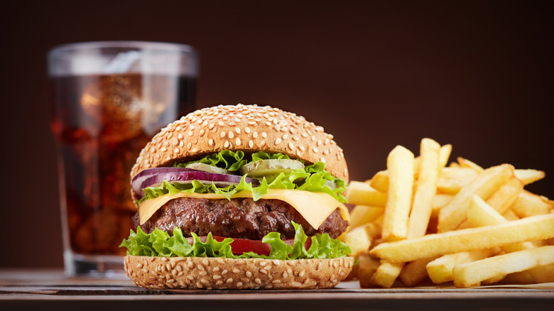 Fast food burger with lettuce, onions, pickles, and cheese, with fries and a soda behind it.