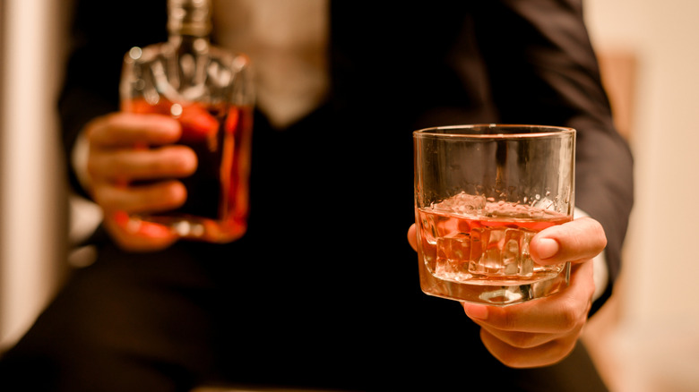 A person's hand is seen holding a glass of whiskey with a finger to indicate its measurement.