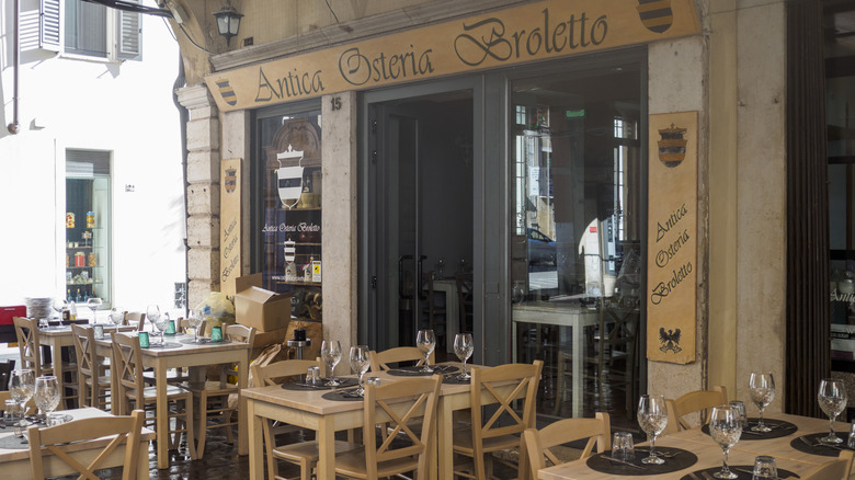 A little osteria has wooden tables topped with wine glasses on a sidewalk patio.