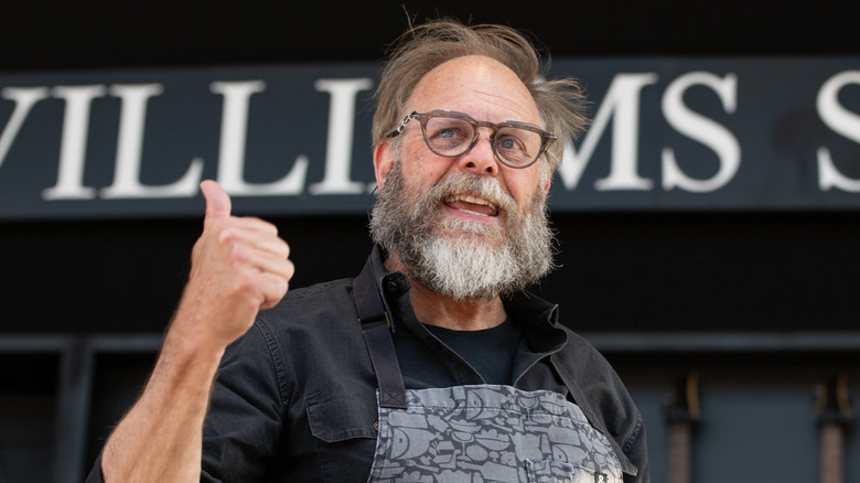 Alton Brown with a long beard in a black shirt and gray apron in front of a wall of guitars and cookware