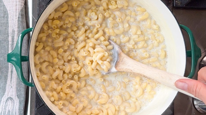 Using wooden spoon to stir macaroni and milk cooking together in large pot on stovetop