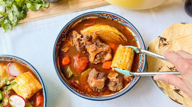 Placing a corn cob into a bowl of Caldo de Res with tongs