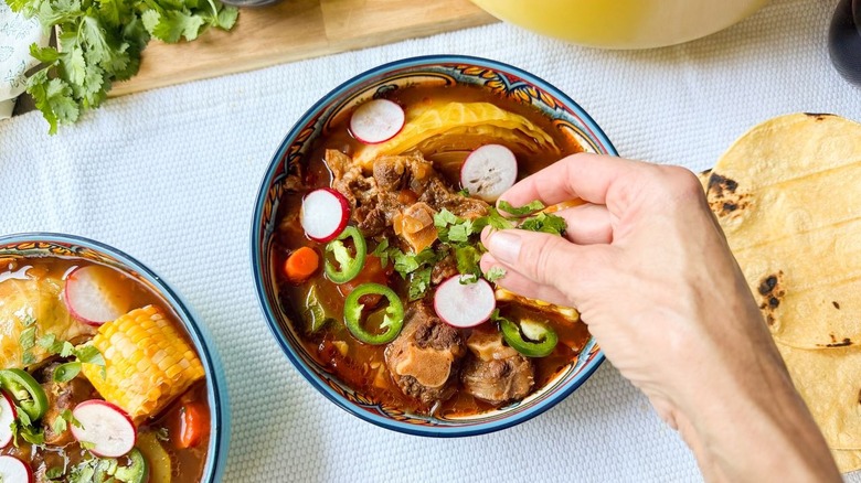 Garnishing bowl of Caldo de Res with sliced radish, jalapeño, and chopped cilantro