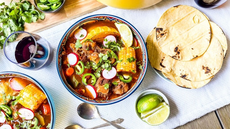 Bowls of Caldo de Res on table with tortillas, lime wedges, spoons, and red wine