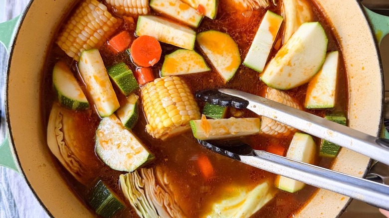 Adding vegetables to Caldo de Res with tongs in Dutch oven