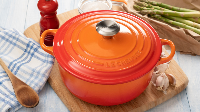Le Creuset lidded pot in flame set on cutting board with wooden spoon and checkered cloth nearby.