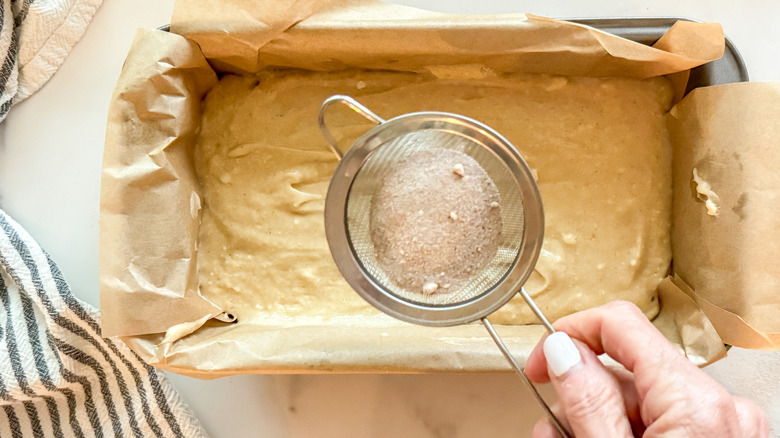 small sieve with cinnamon sugar hovering over unbaked bread in loaf pan