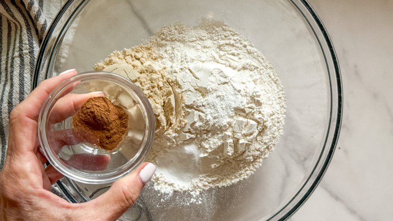 hand adding cinnamon to bowl with flour in it