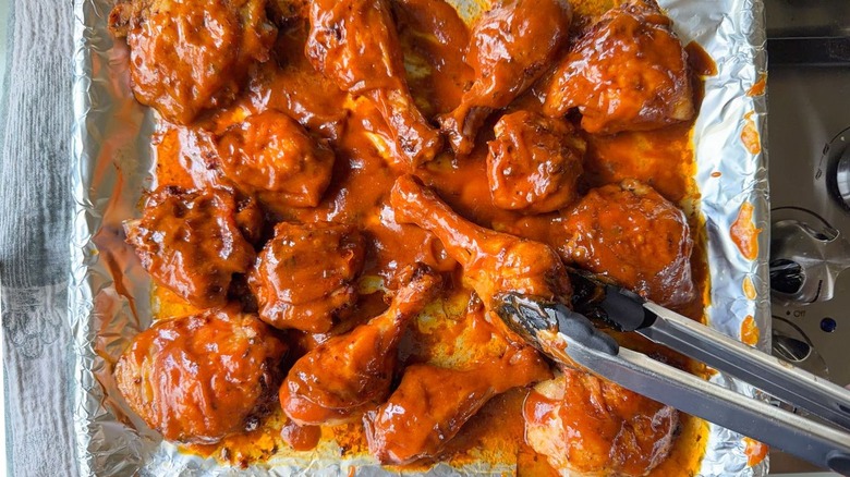 Coating baked chicken legs and thighs with honey bourbon BBQ sauce on foil-line baking pan with tongs