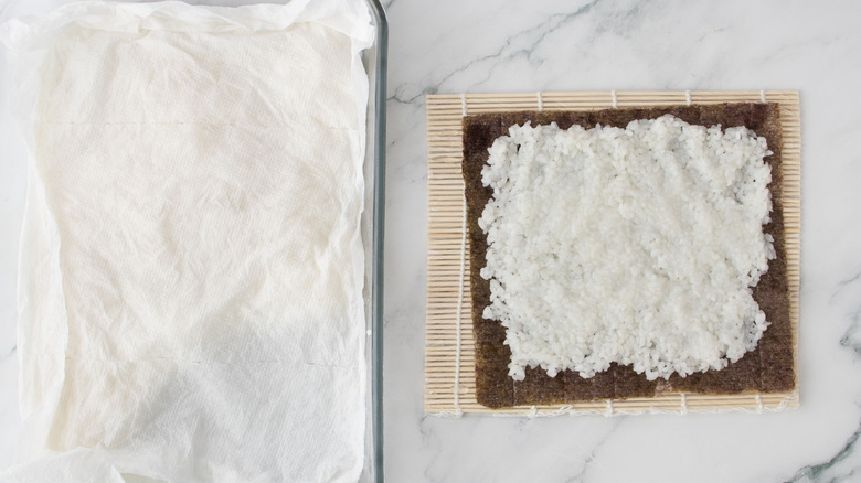 Paper towel-covered casserole dish next to bamboo sushi mat with nori and rice layer