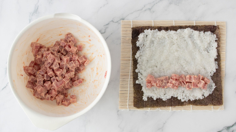 Bowl with cubed tuna next to bamboo sushi mat with nori, rice, and tuna layer