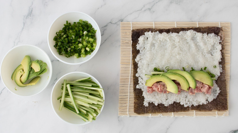 Small bowls of avocado, scallions, and cucumber next to unrolled spicy tuna roll on sushi mat