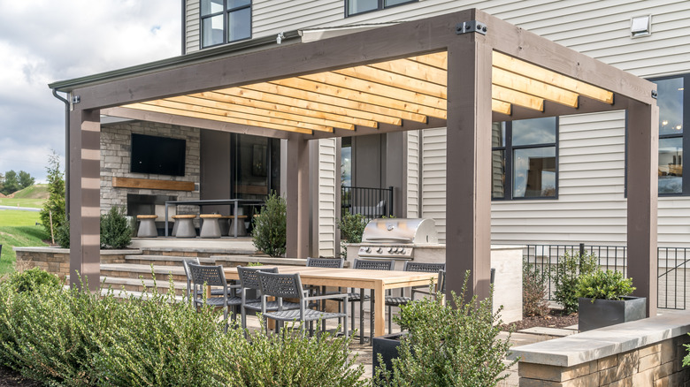 A grill and dining table sit under a pergola outside a large modern house