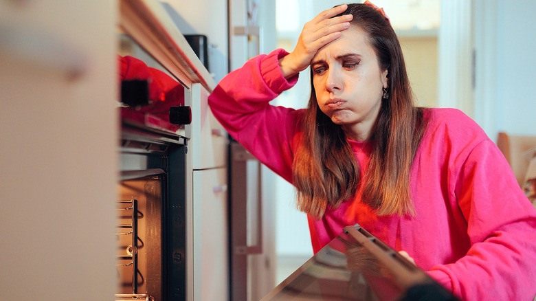 Women in a bright pink, long-sleeve shirt holding her forehead in distress while looking inside an oven.