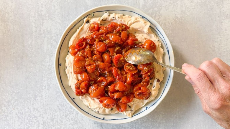 Spreading warm roasted cherry tomatoes on top of BLT dip base in shallow bowl