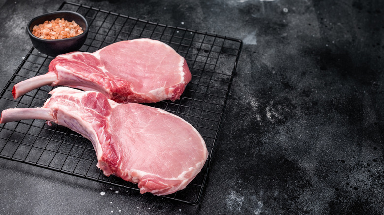 A pair of uncooked pork chops are arranged on a wire rack with a pot of salt.