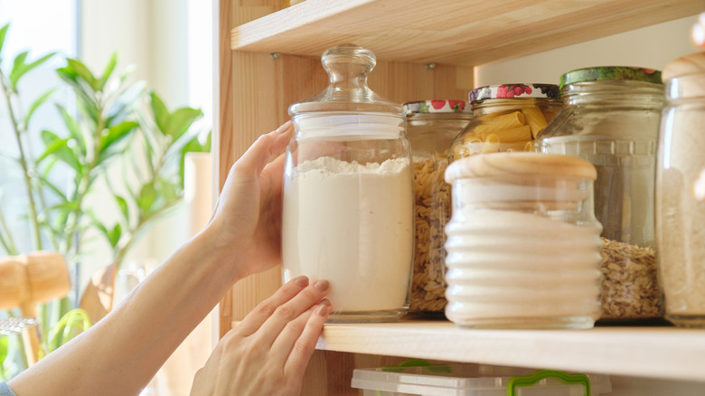 A person's hand is seen grabbing a glass jar of flour from a shelf.