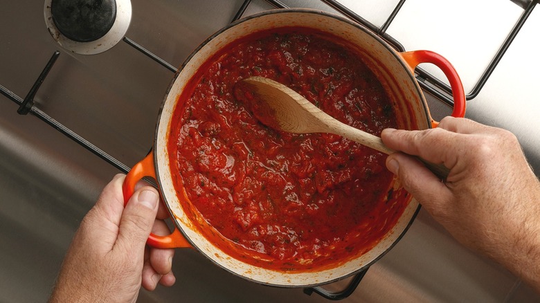 Hands stirring red pasta sauce on a stovetop with a wooden spoon.