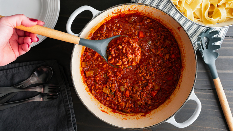 A hand holding a wood and silicone spoon full of meat sauce over a full pot of it, next to a bowl of pasta