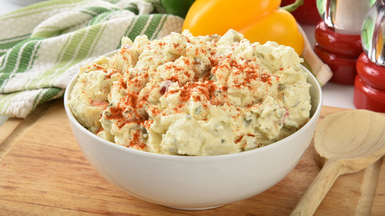 A bowl of mayo and mustard potato salad, topped with paprika on a wooden cutting board, a wooden spoon to the side, a tea towel in the background along with bell peppers.