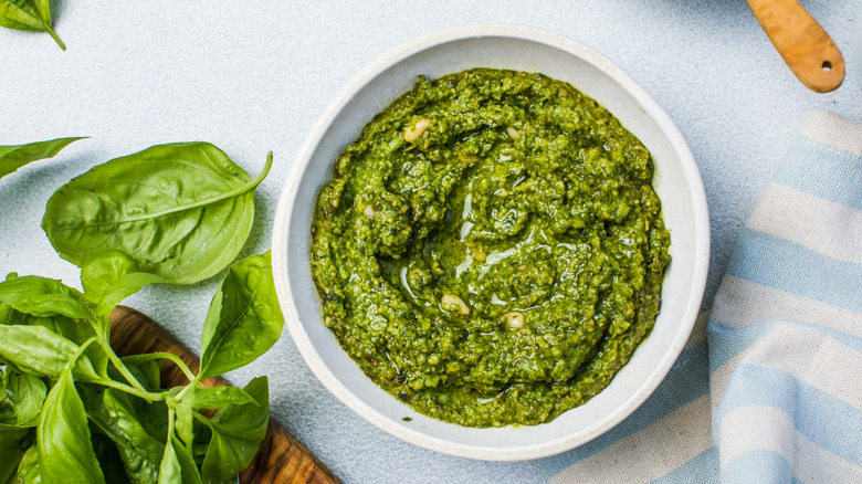 Green pesto in a white bowl beside basil leaves.