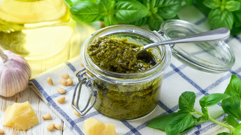 A jar of vibrant green pesto on a kitchen towel surrounded by basil, garlic, and chunks of Parmesan