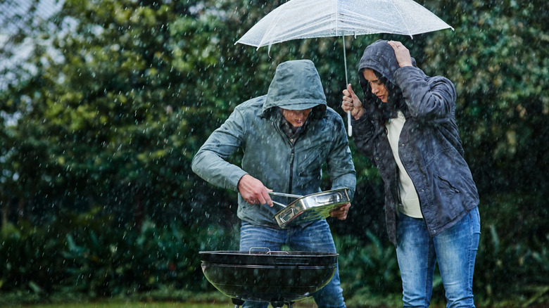 A man in a raincoat and blue jeans uses tongs to pull food off the grill while a worried looking woman hold an umbrella over both of them in the rain.