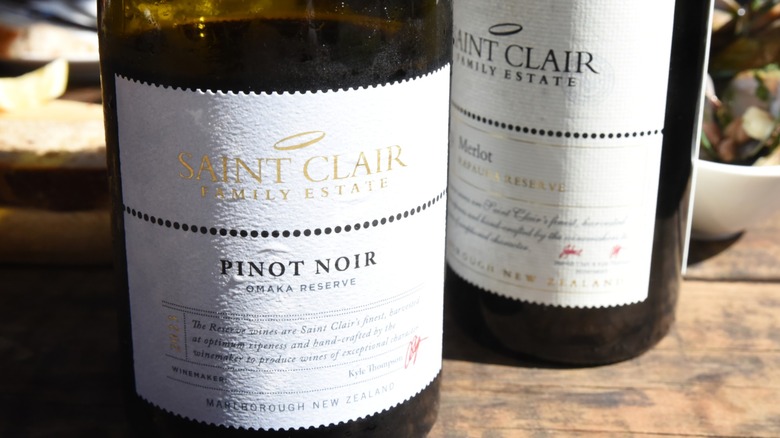 A bottle of pinot noir and a bottle of merlot appear on a rustic wooden table.