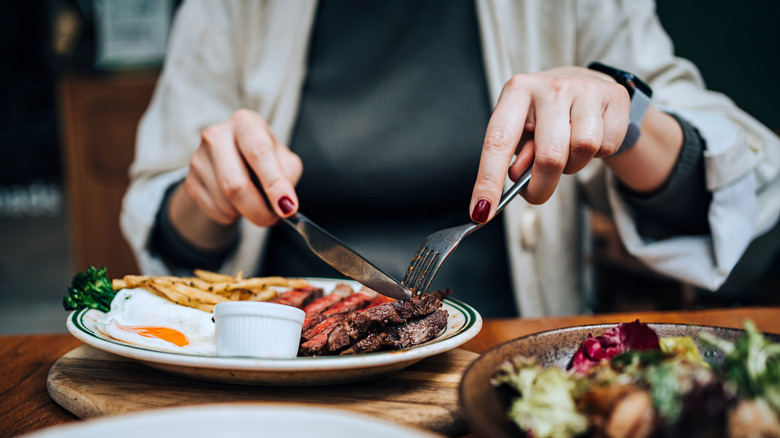 A person's hands are seen cutting a steak with a knife and fork in a restaurant setting.
