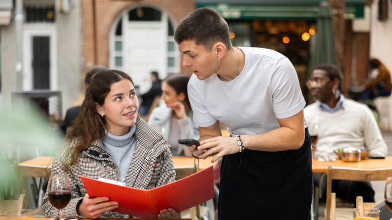 A restaurant patron holds a menu while speaking to a server.
