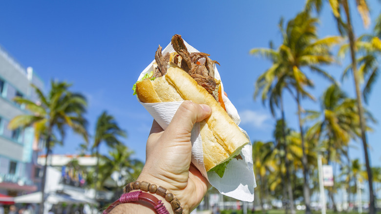 Person holding a Cuban sandwich against palm trees
