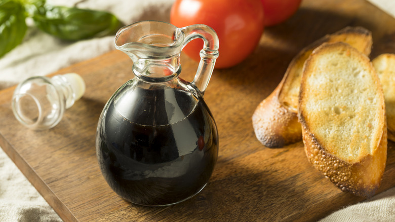 A jug of balsamic vinegar on a wooden board with slices of bread and tomatoes