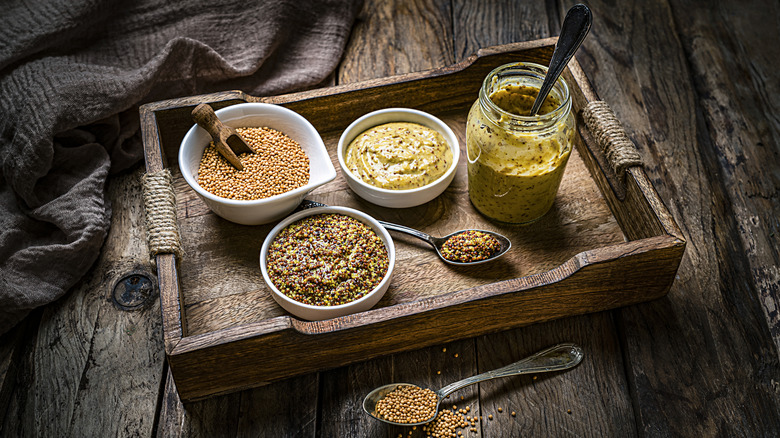 Array of mustards seeds and dips in bowls and jars with spoons on a wooden tray