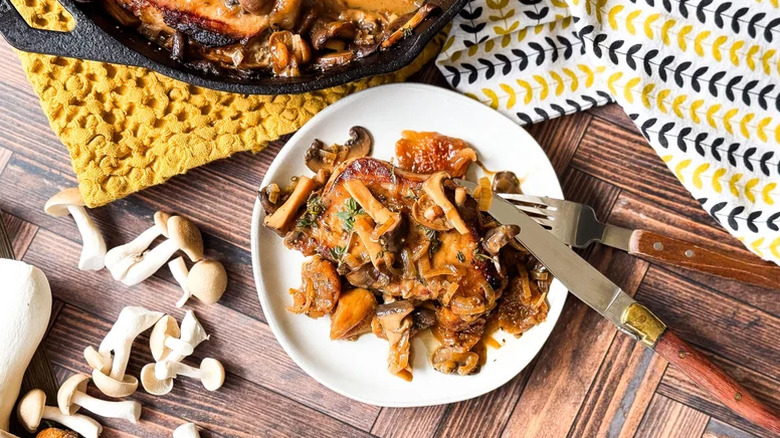 A serving of pork chops covered in mushrooms on a white plate with fork and knife.