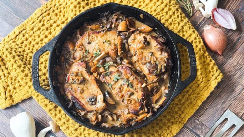 Smothered pork chops in a cast iron pan on a yellow dish towel.
