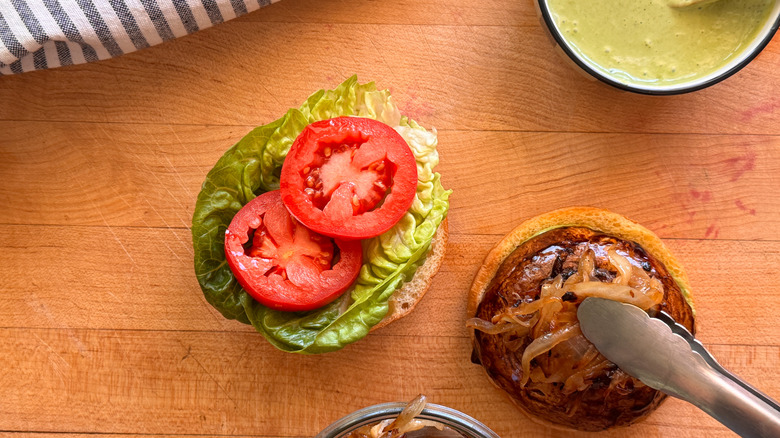 Open faced burger with tomatoes, lettuce, and tongs placing onions
