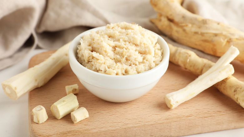 Bowl of grated horseradish on a wooden board with whole, peeled horseradish on the side